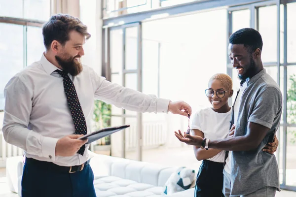 Side view of cheerful bearded formal male employee holding clipboard and giving house key to excited black couple in modern outfit