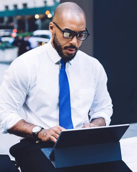 Concentrated bald and bearded African American businessman in white shirt with blue tie and eyeglasses sitting on bench and hardworking on tablet