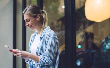 Moda kıyafetleri içinde neşeli sarışın bir kadın şehrin dışındaki kafenin penceresinin yanındaki akıllı telefondan oturmuş mesajlaşıyor.