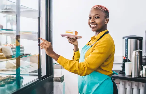 Side view of young smiling African American female worker of pastry shop pointing at cake display fridge and holding plate with piece of cake while inviting for trying delicious sweets