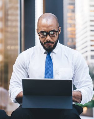 Kendine güvenen kel ve sakallı Afrikalı Amerikalı girişimci resmi olarak kravat ve gözlük takıyor ve bulanık arka planda tablet kullanıyor.