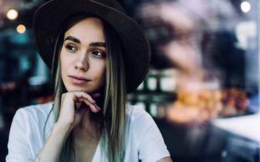 Tropikal şapkalı, beyaz gömlekli, sol tarafa bakan, çene kenarında el ele tutuşan arka planda bulanık bir kafede oturan genç kadının cam manzarası.