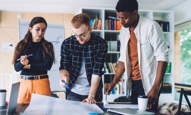 Çağdaş ofiste masanın etrafında dikilip mimari proje üzerinde çalışırken taslak hazırlayan ve birbirleriyle konuşan çok ırklı gençler