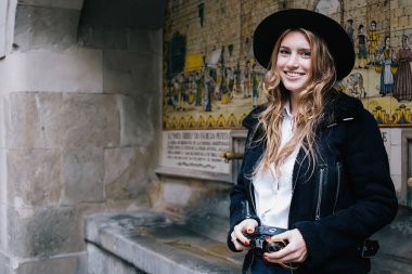 Şapkalı ve kameralı neşeli genç bayan turist şehir gezisi sırasında duvarda durup gülümsüyor ve tarihi resimler çiziyor.