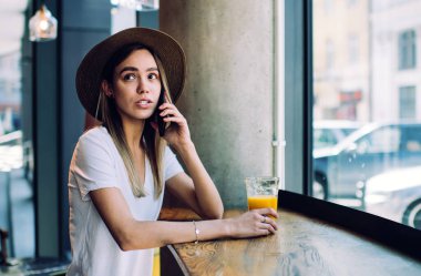 Elinde meyve suyu bardağıyla akıllı telefondan konuşan ve restoranın penceresinin yanındaki masada otururken yüzünü çeviren güzel genç bayanın yan görüntüsü.