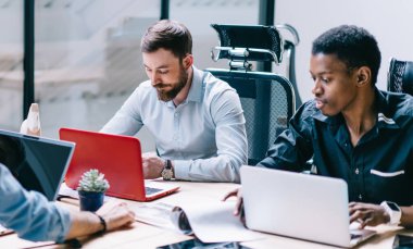 Çeşitli yoğun yoğun yoğun iş ortakları iş kıyafetleri giyiyorlar. Gündüz vakti büyük bir masada otururken dizüstü bilgisayarlar kullanıyorlar.