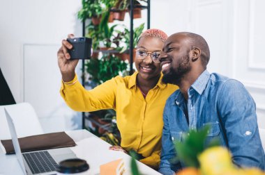 Heyecanlı Afro-Amerikalı genç bir kadın, günlük kıyafetler ve gözlüklerle fotoğraf çekiyor. Kamerayla modern kafede içerikli siyah bir erkekle rahatlıyor.