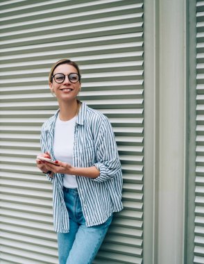 Gündelik giysiler içinde neşeli genç bir kadın ve modaya uygun gözlükler takmış akıllı telefon kullanırken ve sokakta pencereye yaslanırken gülümsüyor ve uzağa bakıyor.