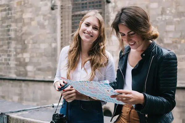 Tourist smiling women in denim and casual wear sitting on stone fence in historical location in city and reading map