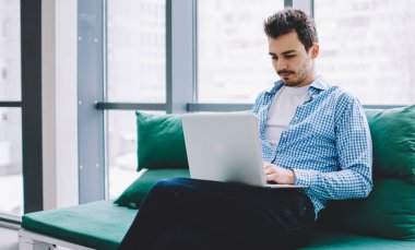 Gelişigüzel giyinen, bacak bacak üstüne atmış, modern dizüstü bilgisayarla iş yerinde bankta oturan genç bir adam.