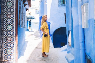 Sarı elbiseli, omzunda fotoğraf makinesi olan genç bir kadın dar bir sokakta mavi duvarlı eski taş binalarla Fas 'ı gezerken görülüyor.