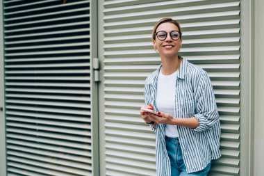 Gündelik giysiler içinde neşeli genç bir kadın ve modaya uygun gözlükler takmış akıllı telefon kullanırken ve sokakta pencereye yaslanırken gülümsüyor ve uzağa bakıyor.