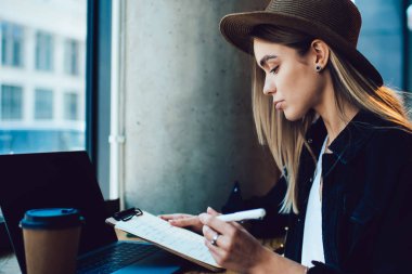 Kafe masasında dizüstü bilgisayarı ve kahveyle otururken panoda notları kontrol eden modaya uygun şapkalı ciddi genç bir kadının yan görüntüsü.