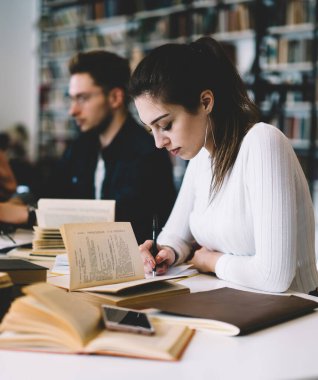 Üniversite projesinde çalışan, rahat giyinen, edebiyat okuyan, not defterinde not tutan kütüphanedeki masada oturan genç bir kadın.