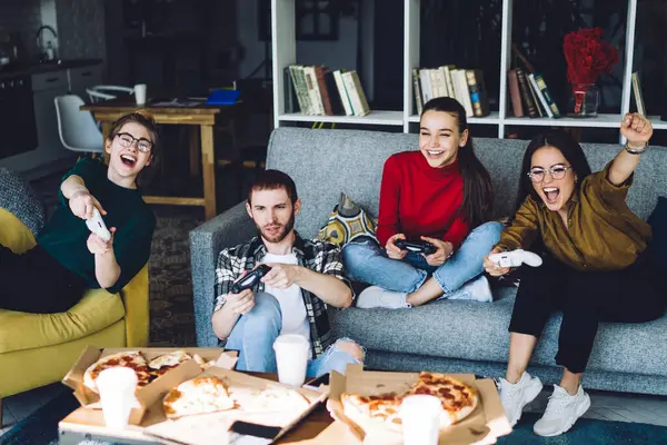 Smiling young gamers playing console with game pad and raising hand in honor of victory eating pizza sitting on sofa in living room with book shelves