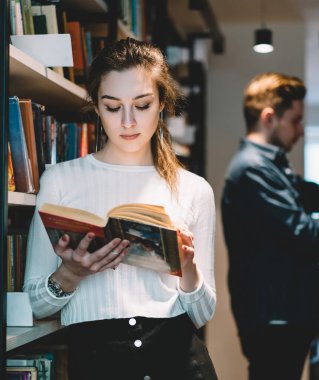 Moda kıyafetleri içinde konsantre kadın müşteri ilginç kitapları kontrol ederken şık bir kitapçıda arka planda erkek alışverişçiyle kitap raflarının arasında duruyor.