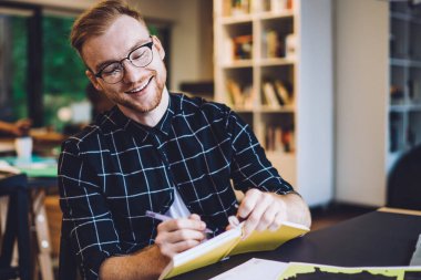 Genç, neşeli hippi sakallı, gözlüklü, siyah beyaz tişörtlü, üniversite kütüphanesinde okurken not defterine yazan kızıl saçlı adam.