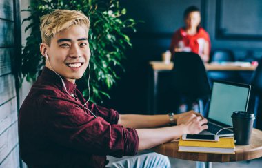 İyimser gülümseyen, kulaklıklı, kulaklık takan, iş yerindeki masada oturup kameraya bakarak dizüstü bilgisayar kullanan Asyalı adamın yan görüntüsü.