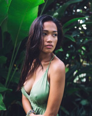 Uzun saçlı, güzel zeytin tenli, açık saçık kesim elbiseler giyen, tropikal bahçede dikilen ve gözlerini kaçıran Asyalı kadının yan görüntüsü.