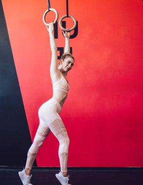 Renkli modern spor salonunda siyah ve kırmızı duvarın yanında uzanan spor ekipmanlarına karşı bej renkli etek giyen, gülümseyen ve jimnastik halkaları üzerinde egzersiz yapan formda bir sporcu.