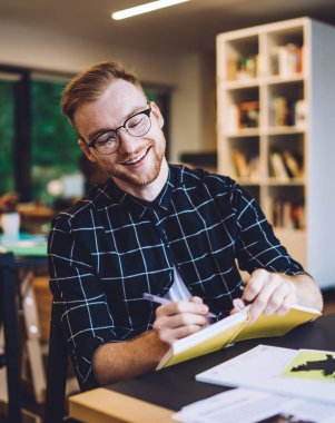 Genç, neşeli hippi sakallı, gözlüklü, siyah beyaz tişörtlü, üniversite kütüphanesinde okurken not defterine yazan kızıl saçlı adam.