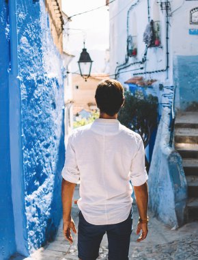 Fas 'ın yaşlı bir kasabasında güneşli bir günde dar sokaktaki eski evlerin önünde duran beyaz gömlekli, isimsiz bir adamın arka görüntüsü.