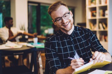 Genç, neşeli hippi sakallı, gözlüklü, siyah beyaz tişörtlü, üniversite kütüphanesinde okurken not defterine yazan kızıl saçlı adam.