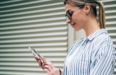 Beyaz tişörtlü, çizgili tişörtlü, gözlüklü, kırmızı tırnaklı, akıllı telefondan kapalı jaluzinin yanındaki videoyu izleyen şık bir kadının yan görüntüsü.