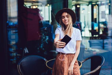 Şık giyinmiş, şapkalı güzel bir kadın akıllı telefondan selfie çekerken sokak kafesindeki masaya yaslanıp kulaklıklarla müzik dinliyor ve gülümsüyor.