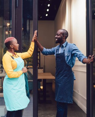 Üniformalı neşeli iş ortakları birbirlerine bakıp kendi kafenin girişinde dururken beşlik çakıyorlar ve başarıyı kutluyorlar.