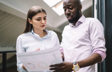 Gündelik giysili Afrikalı Amerikalı şüpheli adam modern ofisteki genç bir bayanın projesine bakıyor.