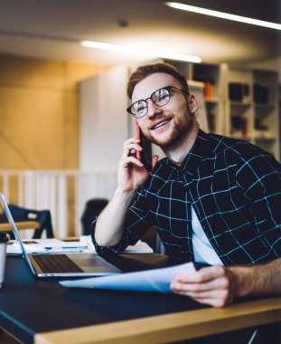 Elinde kağıt olan genç adam bilgisayarla masada otururken gülümsüyor ve düşünüyor ve çağdaş çalışma alanında akıllı telefon konuşmaları yapıyor.