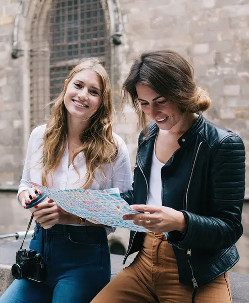 Tourist smiling women in denim and casual wear sitting on stone fence in historical location in city and reading map