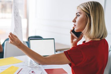 Ofiste açık dizüstü bilgisayarla cep telefonuyla konuşurken, belgede çalışan nitelikli bir yöneticinin yan görünümü