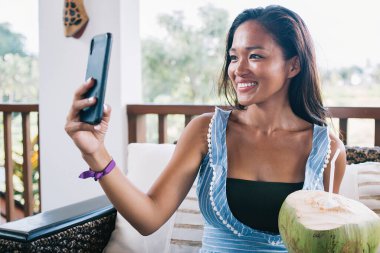 Mavi elbiseli çekici bir Asyalı kadın akıllı telefonuyla selfie çekerken kanepede oturuyor ve kırsal alanda bulanıklığa karşı balkonda serinliyor.