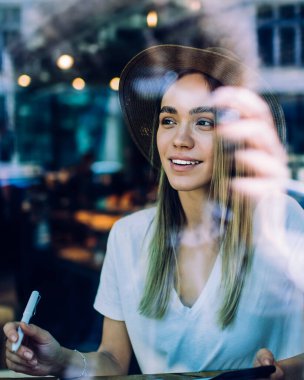 Camdan manzaralı, tropikal şapkalı ve beyaz gömlekli genç bir kadın bulanık arka planda, kafede otururken sol tarafa bakıyor.