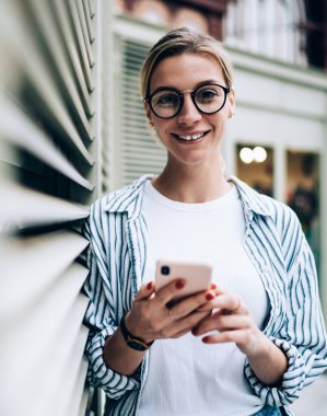 Neşeli sarışın kadın günlük kıyafetler giyiyor ve moda gözlükler takıyor. Gülümsüyor ve kameraya bakıyor. Duvara yaslanıyor ve akıllı telefonlara bakıyor.