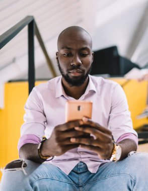Alttan, akıllı telefon ekranına odaklanan, kolejde merdivenlerde oturan, konsantre olmuş genç, kel bir erkekten.