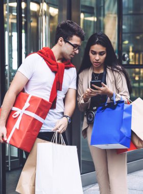 Alışverişten sonra alışveriş merkezinin girişinde duran ve akıllı telefon kullanan çok ırklı bir çift.