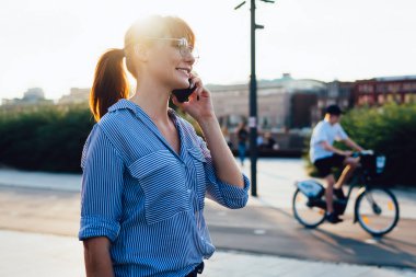 Şehir caddesinde cep telefonuyla konuşurken gülümseyen günlük kıyafet ve gözlüklü bir kadının yan görüntüsü