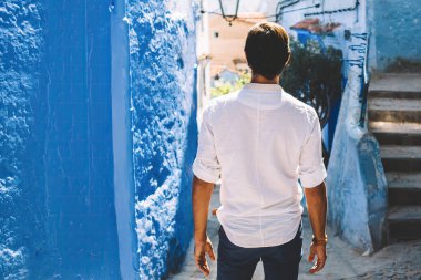 Fas 'ın yaşlı bir kasabasında güneşli bir günde dar sokaktaki eski evlerin önünde duran beyaz gömlekli, isimsiz bir adamın arka görüntüsü.