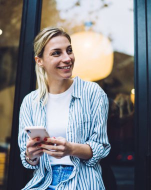 Moda kıyafetleri içinde neşeli, şık, sarı saçlı kadın. Pencerenin yanında oturmuş, akıllı telefonuyla gülümsüyor ve mesajlaşırken başka tarafa bakıyor.