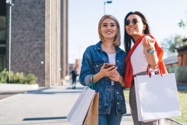 Neşeli kadın kamerayı işaret ediyor. Güneşli bir günde kot pantolon giymiş, alışveriş torbalarıyla rahatlıyor.