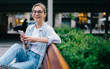 Yuvarlak gözlüklü ve akıllı telefonlu, yeşil çalılığın yakınındaki kahverengi bankta dinlenen, bulanık sokak arka planında, günlük kıyafetli, sarışın bir kadın.