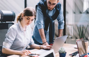 Masaüstünde oturan ve akıllı telefon kullanan konsantre kadın yönetici, beyaz erkek çalışanın yanında durup bilgi bekliyor. İki iş arkadaşı iş çözümlerini analiz etme ve arama