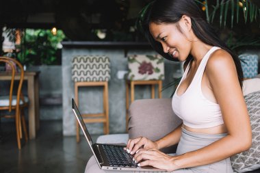 Kafe kanepesinde yaratıcı bir iş yaparken Asya 'lı kadın bilgisayarın zevkle sörf yaparken gülümsemesinin yan görüntüsü.