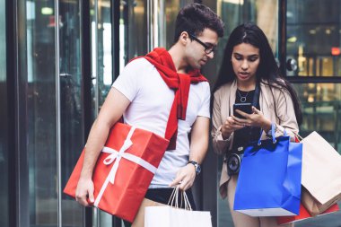 Alışverişten sonra alışveriş merkezinin girişinde duran ve akıllı telefon kullanan çok ırklı bir çift.