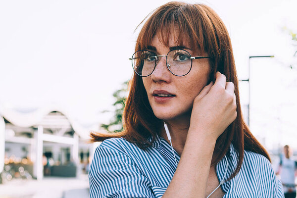 Привлекательная рыжая женщина в очках, надевающая наушники, слушающая плейлист через мобильный телефон на улице, хипстерская девушка Мелломан наслаждается онлайн-радио через приложение на смартфоне и электронный аксессуар