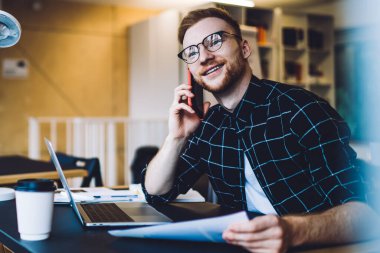 Elinde kağıt olan genç adam bilgisayarla masada otururken gülümsüyor ve düşünüyor ve çağdaş çalışma alanında akıllı telefon konuşmaları yapıyor.