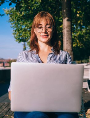 Neşeli kızıl saçlı kadın görüş koruması için serbest çalışıyor video seyrediyor ve laptopta haber okuyor güneşli bir günde dışarıda oturuyor, kadın dijital göçebe şehirde uzaktan çalışıyor.